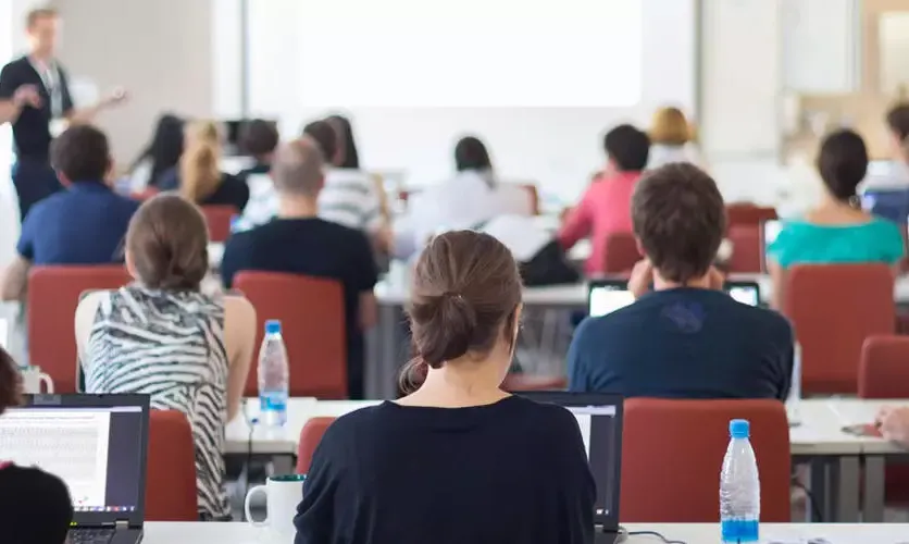 Photo of a classroom from the back where you can see the backs of students' heads and the professor at the front of the room.